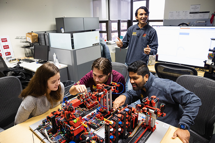 Three information systems students work together on a small machine while a faculty member watches their progress.