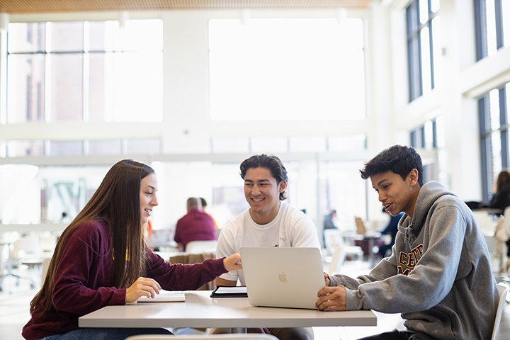 Three students sit at a table looking at a laptop computer in a brightly lit room.