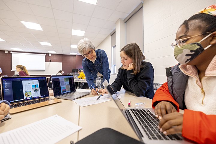 A female professor with white hair and glasses assists two students who sit at a table during Spanish class.