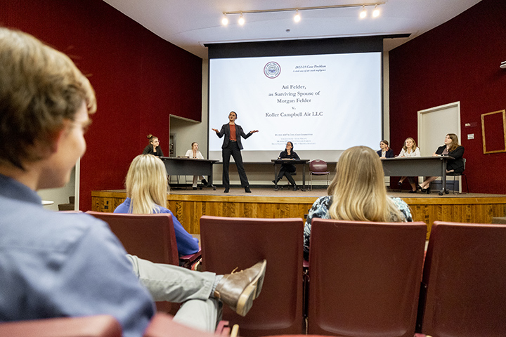 Students sit in an auditorium while six students and a faculty member stand on stage conducting a mock trial.