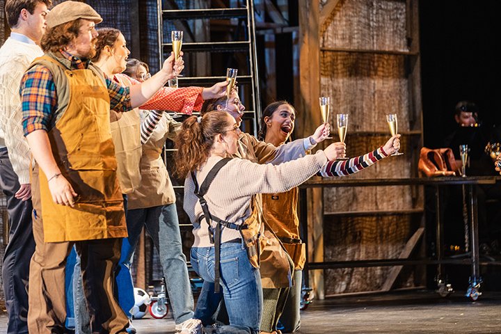 Student cast members stand on stage while singing and holding champagne flutes.