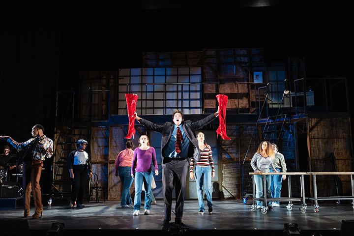 A student actor holds two large, red boots in his hands while a his cast members sing in the background.