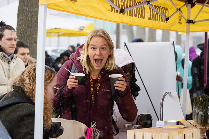 A blonde student holds a cup of soup in each hand while smiling at the camera.