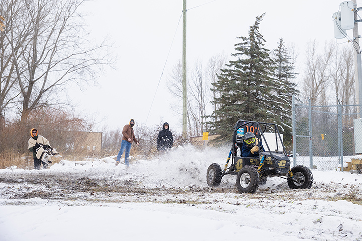 Three students watch as a Baja race car with a CMU logo on its side door races through the snow.