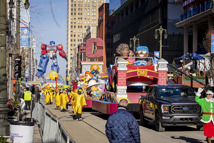 A black pickup truck pulls the CMU parade float as marchers in yellow graduation gowns and hats walk alongside.