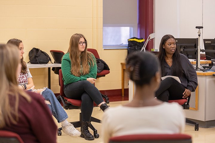 Five students sit in chairs in a group while having a conversation.