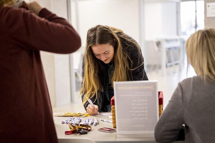 A female student signs a sheet of paper at a table while two College of Business staff members wait to give advice on how best to study during exam week.