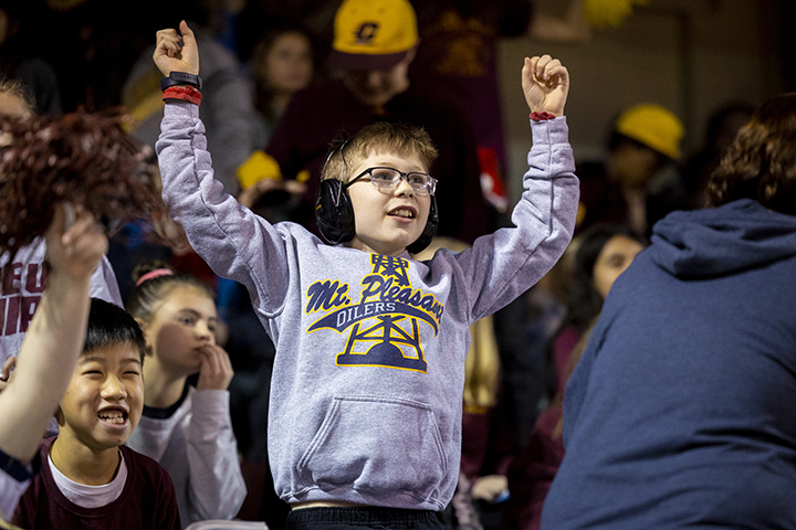 A male elementary-aged student wearing glasses, headphones and a Mt. Pleasant Oilers sweatshirt raises his hands and cheers.