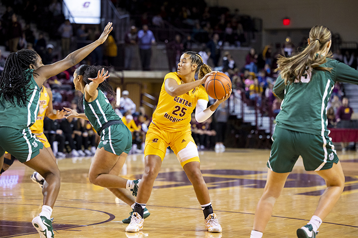 A CMU women's basketball player in a yellow uniform looks to pass to a teammate underneath the hoop as a Cleveland State University player in a green uniform guards her.