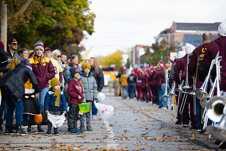 A group of children stand on the side of the road watching the marching band walk past.
