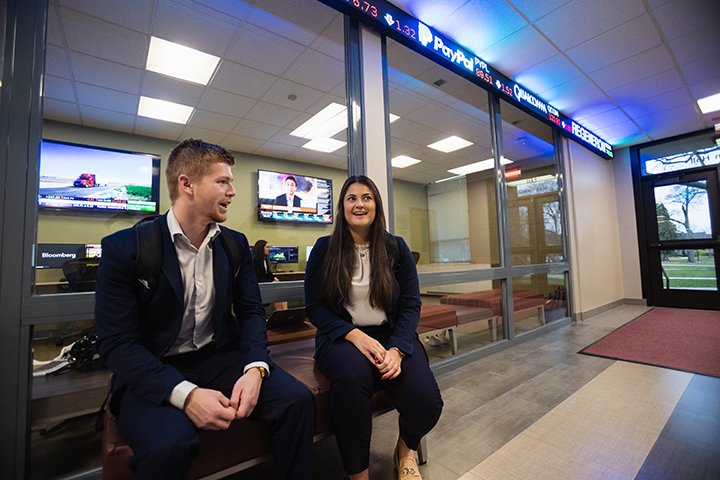 A male and female student sit on a stool in the hallway outside a classroom that features glass walls and TVs on its walls.