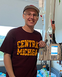 A young man in a Central Michigan shirt and hat smiles while standing in a hospital room.