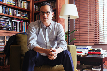 An Asian-American man wearing a collared shirt and blue jeans sits on a mustard-colored chair in a room with a book case filled with books in the background.