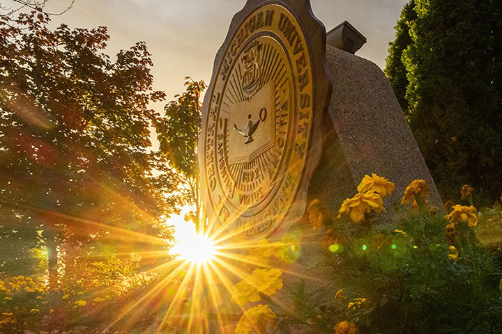 The sunrise peeks through the trees with the CMU seal and flowers in the foreground.