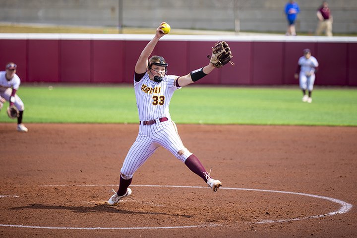 A CMU softball pitcher wearing a white uniform and a protective mask winds up to make a pitch.