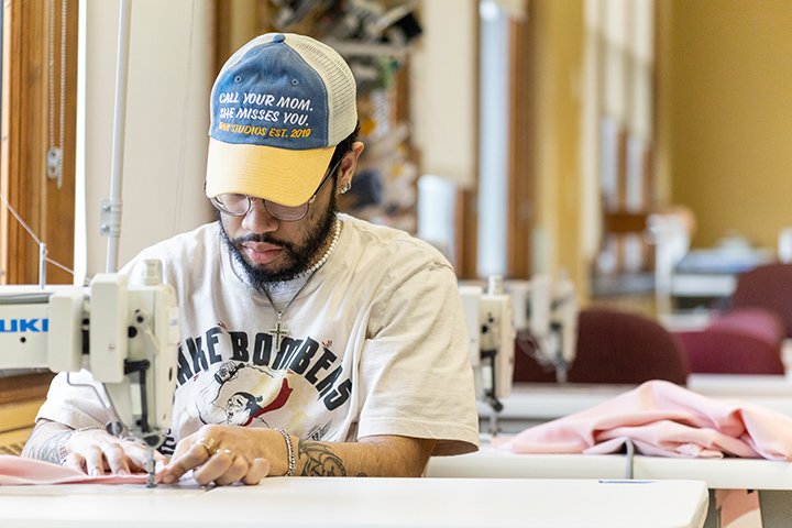 A college student in a hat and glasses uses a sewing machine.