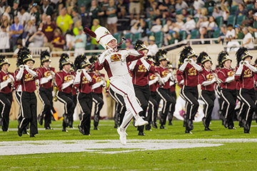 A CMU drum major wearing a white CMU uniform and large white hat leads the CMU marching band onto the field at Spartan Stadium as a large crowd looks on from the stands.