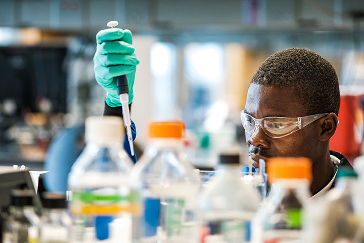 A man wearing safety glasses and holding a pipette researches tuberculosis in a chemistry lab.