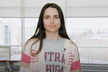 A woman with long brown hair wearing a Central Michigan University shirt and sweater speaks during a YouTube video.
