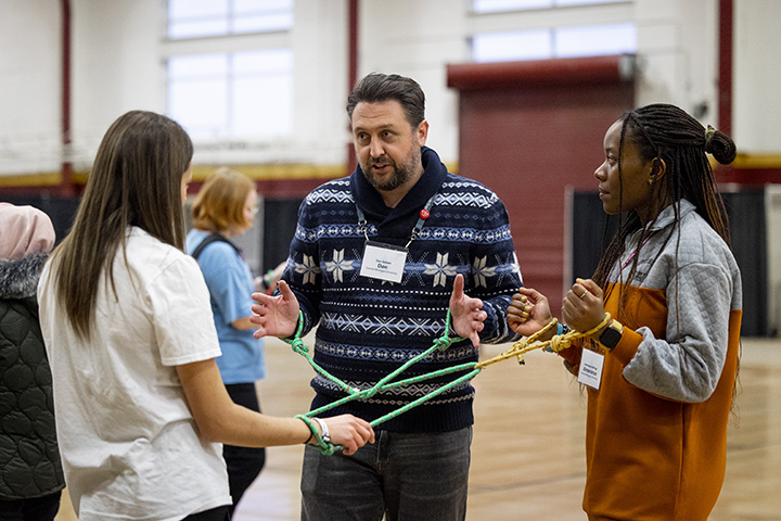 A man with a beard and two female college students have their wrists tied together with rope as part of a team-building exercise.