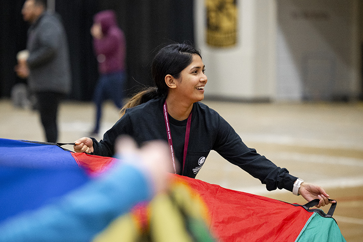 A female college student standing in a gymnasium smiles while holding the edge of parachute as part of team-building exercise.