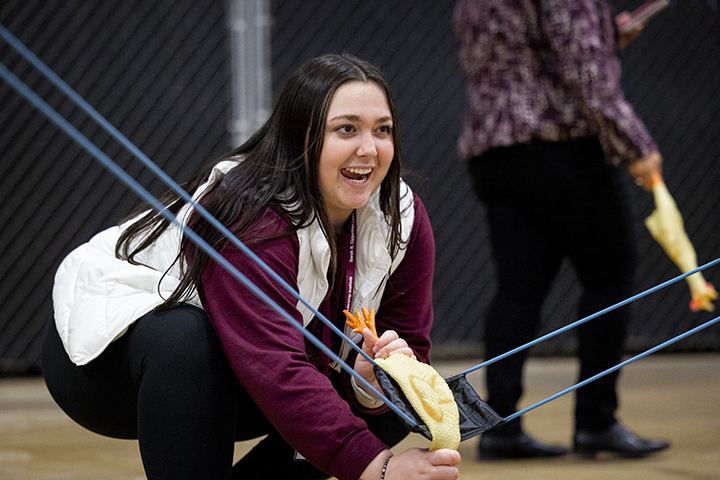 A female college student smiles while aiming to fling a rubber chicken from a slingshot.