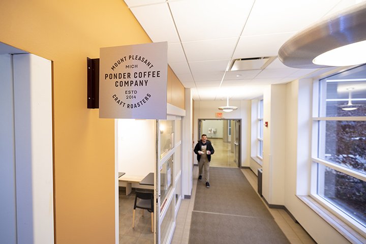 A sign on a yellow wall inside the Health Professionals Building reads “Ponder Coffee Company, Mount Pleasant, Michigan, Craft Roasters, Established 2014” as a male customer walks past the entrance with a coffee in his hand.