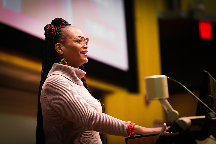 An African-American woman in a dark pink sweater sandglasses stands at a podium and smiles.