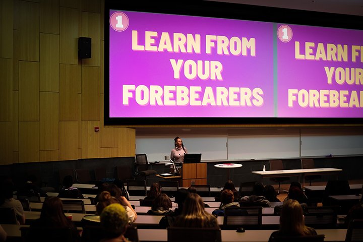A large group of students sit in an auditorium listening to a women in the front of the room as the words