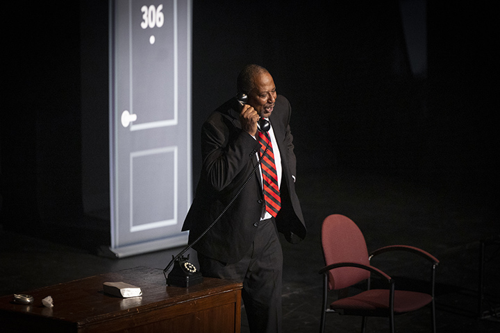 An African-American man in a black suit and red and black tie talks on a phone while performing a one-man show on a stage.