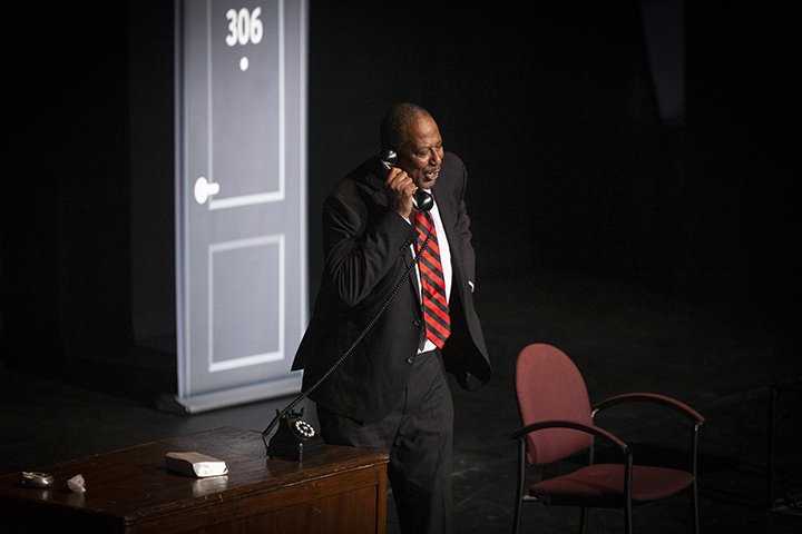 An African-American man in a black suit and red and black tie talks on a phone while performing a one-man show on a stage.