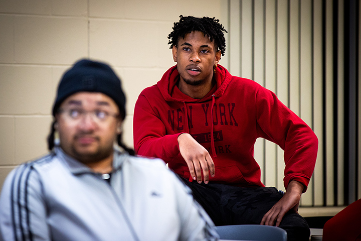A student in a red New York sweatshirt listens intently while another student is out of focus in the foreground.