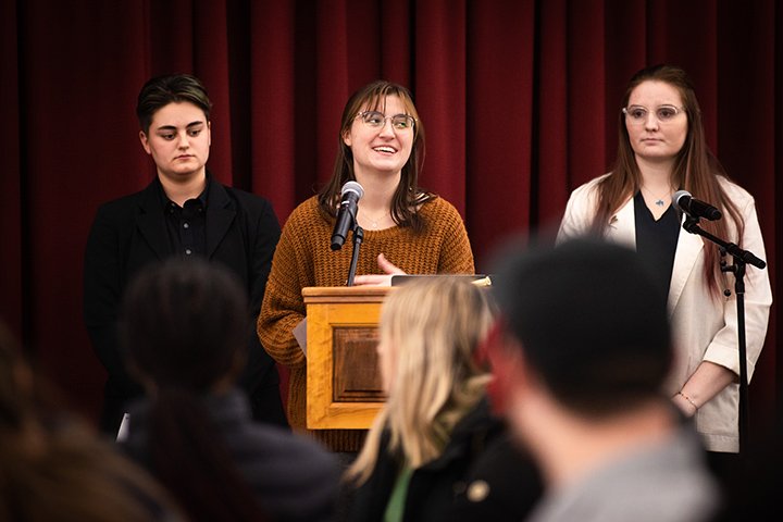 Three students stand at a podium inside the Rotunda Room.