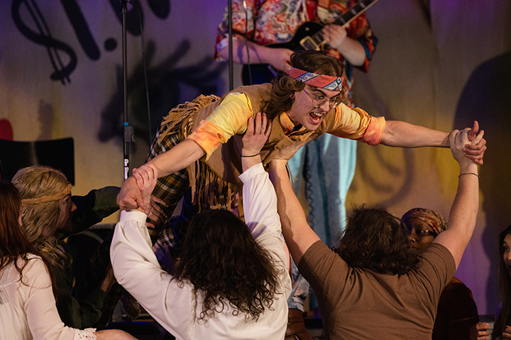 A student actor wearing a bandana and brown, fringe clothing stage-dives onto a small group of people.