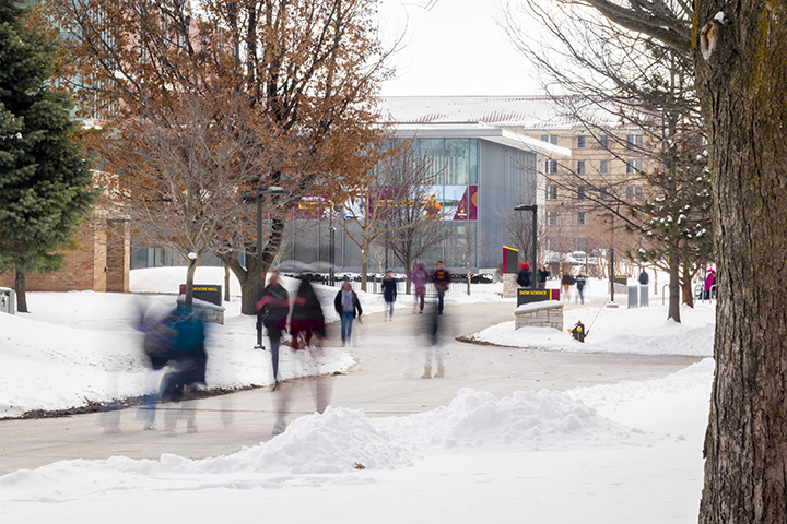 A long-exposure image shows blurry people walking down a sidewalk surrounded by snow as the Dow Science Building stands tall in focus in the background.