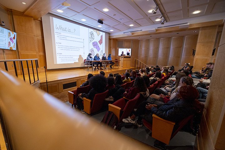 Students sit in an auditorium watch and listen intently as black faculty and leaders sit at a table on stage leading a conversation.