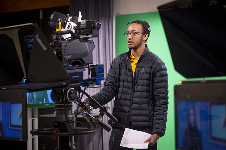 A student in a black coat and glasses runs a television camera in Moore Hall's television studio.