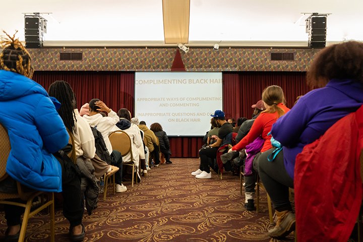 Students sit in chairs in a large room facing a projector display screen with the words 