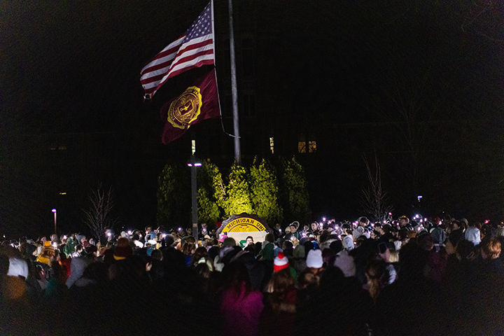 A large American Flag and the CMU seal are lit up at night as hundreds of people gather for candlelight vigil.