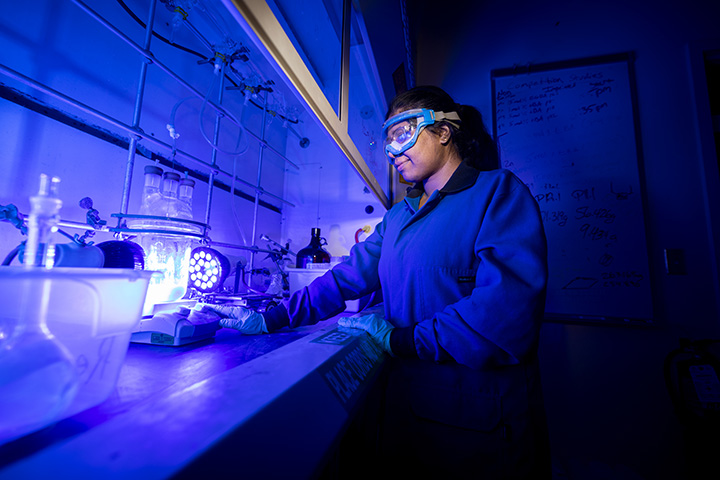 A female student wearing protective eyewear works in a lab that's lit by blue lights.