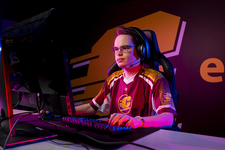 A male student wearing glasses and a CMU-branded maroon jersey sits at a computer playing video games.