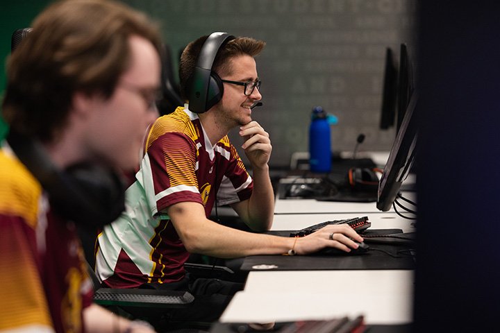 A male student wearing glasses smiles while sits at a computer and playing video games.