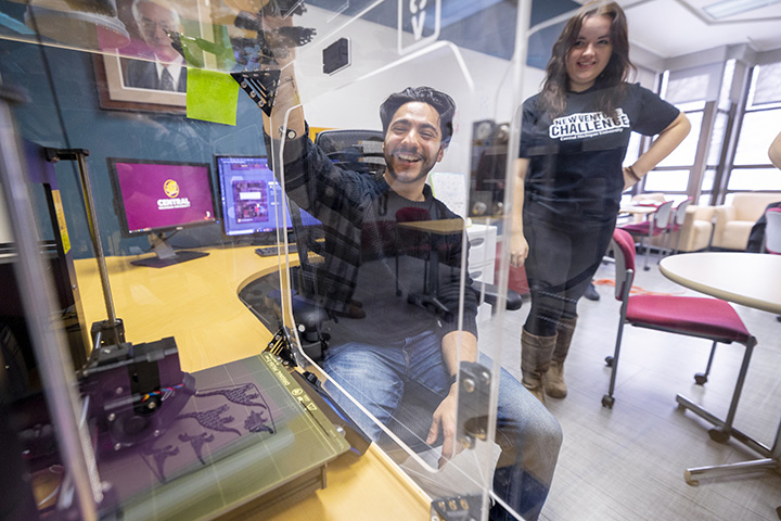 A male student sits in front of a 3D printer smiling while a female student smiles and looks on from the background.