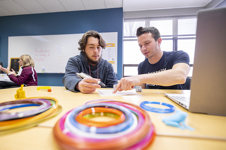 Two male students sit at a table brainstorming ideas. On the table sits a laptop and multiple colored spools of wire.