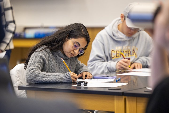 Two students sit at a table inside a classroom writing on pieces of paper.