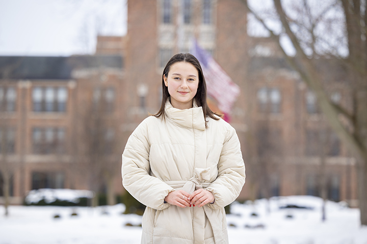 Student Masha Smahliuk wears a cream-colored coat, smiling at the camera while standing in front of Warriner Hall.