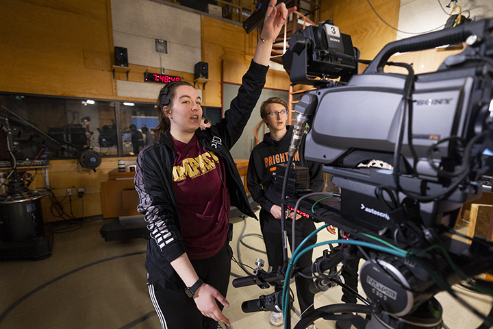Two college students stand next to a large television camera inside a TV studio.