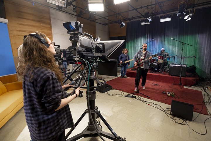 A college student wearing a headset controls a TV camera while a three person band plays inside a studio.