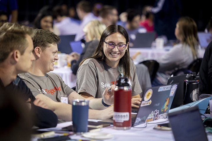 One male student and one female student, both wearing light brown t-shirts, smile at each other during the ERPsim competition.