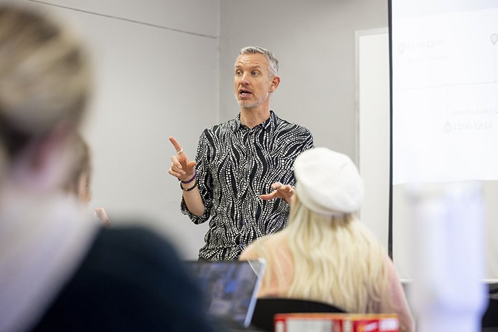 A gray-haired faculty member wearing a black and silver shirt stands in front of a classroom full of students.